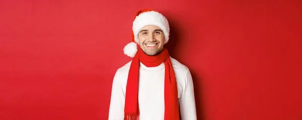 Concepto de vacaciones de invierno, Navidad y estilo de vida. Primer plano del hombre guapo con cerdas, con sombrero de Navidad con bufanda y sonriendo feliz, deseando feliz año nuevo, fondo rojo — Foto de Stock