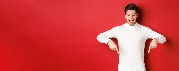 Retrato de homem feliz de boa aparência, apontando os dedos para baixo e olhando para a propaganda com sorriso animado, de pé sobre fundo vermelho — Fotografia de Stock