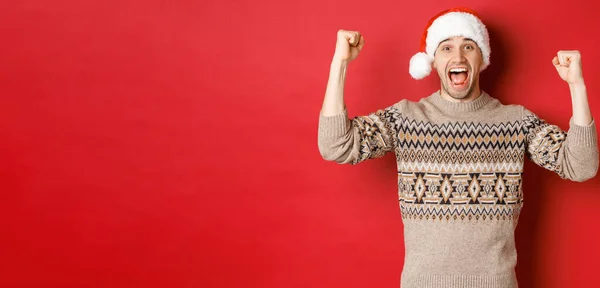 Imagem de homem bonito alegre em jurador e chapéu de Papai Noel, celebrando o ano novo, triunfando ou ganhando algo, levantando as mãos e gritando de alegria, de pé sobre o fundo vermelho — Fotografia de Stock