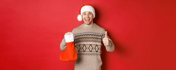 Concept de vacances d'hiver, nouvel an et célébration. Joyeux bel homme en chapeau et pull Père Noël, montrant bas de Noël avec des bonbons et des cadeaux, faire pouces vers le haut — Photo