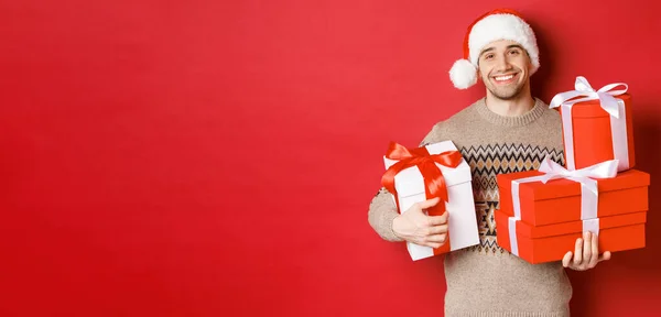 寒假的概念，新年和庆祝活动。可爱的面带微笑的男人为圣诞节准备了礼物，拿着礼物，看着镜头温暖的红色背景 — 图库照片