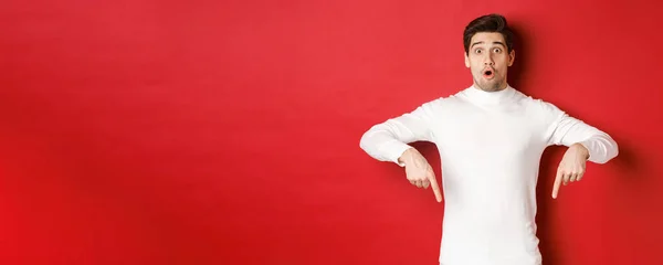 Изображение удивленного привлекательного парня в белом свитере, показывающего объявление, указывающего пальцами вниз и выглядящего изумленным, стоящего на красном фоне — стоковое фото