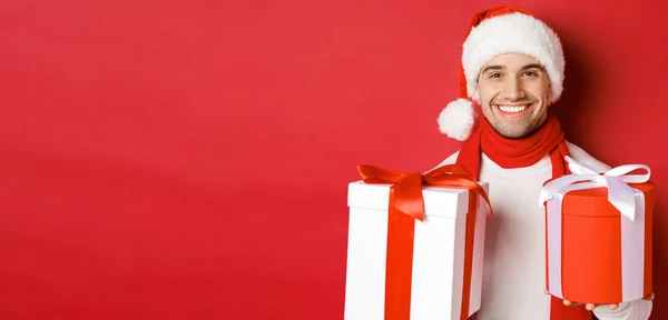 Conceito de férias de inverno, Natal e estilo de vida. Close-up de sorrir cara bonito em santa chapéu e cachecol, desejando feliz ano novo e segurando presentes, de pé com presentes sobre fundo vermelho — Fotografia de Stock