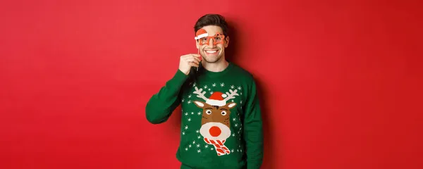 Retrato de belo homem sorridente em camisola de Natal e óculos de festa, celebrando o ano novo e se divertindo, de pé contra o fundo vermelho — Fotografia de Stock