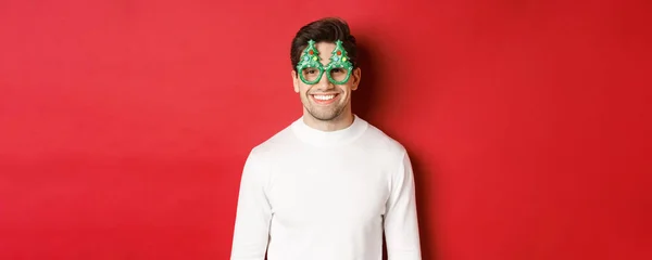 Conceito de Natal, férias de inverno e celebração. Close-up de homem bonito em suéter branco e óculos de festa, desfrutando de ano novo, de pé sobre fundo vermelho — Fotografia de Stock