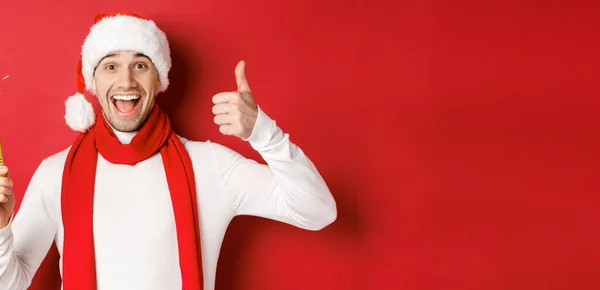 Koncepcja Bożego Narodzenia, ferie zimowe i uroczystości. Przystojny mężczyzna świętuje Nowy Rok i dobrze się bawi, trzyma iskrę i pokazuje kciuk w górze, stoi na czerwonym tle — Zdjęcie stockowe