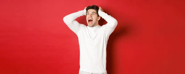 Portret van gefrustreerde man in witte trui, schreeuwend in paniek en kijkend naar de linker bovenhoek, hebben verschrikkelijk probleem, staande over rode achtergrond — Stockfoto