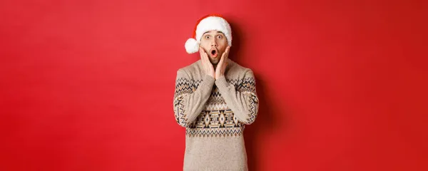 Noel Baba şapkalı, ağzı açık duran ve kırmızı arka planda duran muhteşem Noel teklifini izleyen hayrete düşmüş ve nutku tutulmuş bir adamın görüntüsü. — Stok fotoğraf