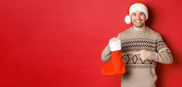 Concept de vacances d'hiver, nouvel an et célébration. bel homme souriant préparé des cadeaux pour les enfants, pointant vers Noël sac bas, debout sur fond rouge — Photo