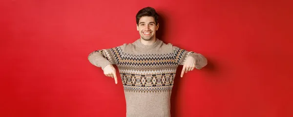 크리스마스, 겨울 휴일, 생활 양식의 개념. xmas 스웨터를 입은 매력적 인 행복 한 남자가 빨간 배경 위에 서 있는 로고를 손가락으로 가리키며 프로모션을 보여 주는 모습 — 스톡 사진