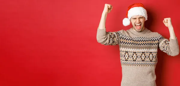 Zufriedener gutaussehender Mann, der sich glücklich und glücklich fühlt, vor Freude schreit und Faust-Pumps macht, Sieg oder Niederlage feiert, ein tolles Weihnachtsgeschenk erhält, im Weihnachtsmann-Hut vor rotem Hintergrund steht — Stockfoto