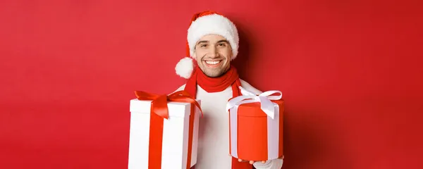 Concepto de vacaciones de invierno, Navidad y estilo de vida. Primer plano de chico guapo sonriente en sombrero de santa y bufanda, deseando feliz año nuevo y la celebración de regalos, de pie con regalos sobre fondo rojo —  Fotos de Stock