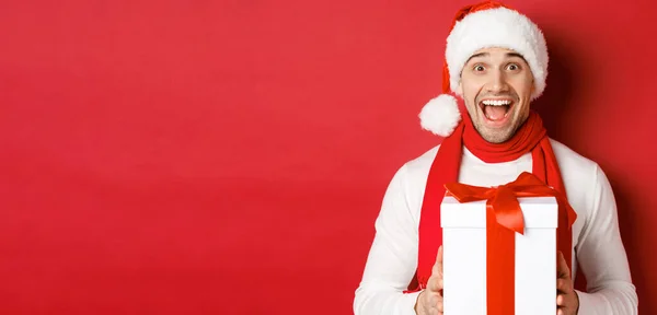 Begrip wintervakantie, kerst en levensstijl. Close-up van opgewonden knappe man in santa jat en sjaal, op zoek verbaasd en het ontvangen van nieuwe jaar cadeau, staande over rode achtergrond — Stockfoto