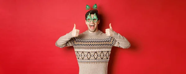 Parti gözlüklü ve Noel süveteri giyen heyecanlı yakışıklı bir adam resmi. Başparmaklarını kaldırıyor, harika yeni yıl kutlamalarını övüyor, kırmızı arka planda duruyor. — Stok fotoğraf
