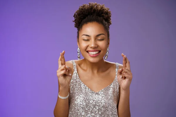 Позитивна щаслива афро-американська молода дівчина, яка святкує день народження, мріючи в елегантній срібній сукні з близькими очима, усміхаючись оптимістичним сном крос-пальців, справджується, стоячи синім фоном. — стокове фото