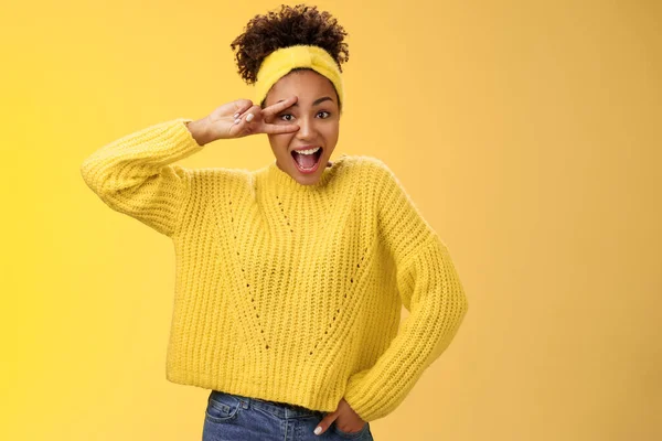 Ενεργητικό γοητευτικό χιλιετή αφροαμερικανή φοιτήτρια σε κίτρινο πουλόβερ κεφαλόδεσμο δείχνουν νίκη χειρονομία ειρήνης αισθάνονται τυχεροί ενθουσιασμένοι απολαμβάνοντας ντίσκο κόμμα χαμογελώντας χαρούμενα, χαρά — Φωτογραφία Αρχείου
