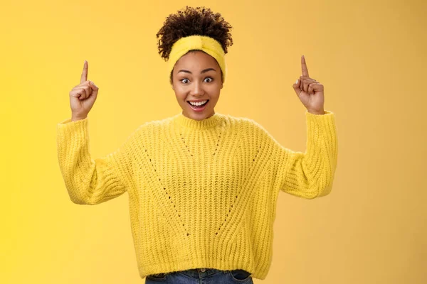 Encantadora mujer afro-americana joven emotiva en diadema de suéter ganando dinero impresionante suma apuntando hacia arriba dedos índice mostrando enlace sorprendido sonriendo ampliamente fondo amarillo — Foto de Stock