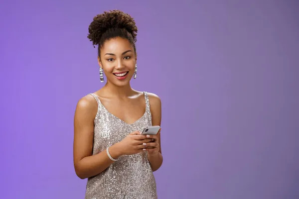 Aantrekkelijke stijlvolle vertrouwen Afrikaans-Amerikaanse vrouw in zilver glanzende jurk met smartphone messaging scrollen internet gadget op zoek gelukkig gelukkig camera staande blauwe achtergrond verheugen — Stockfoto