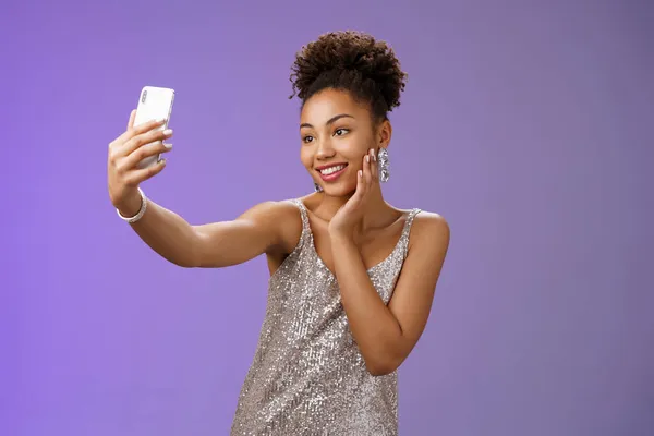 Populaire stijlvolle Afrikaans-Amerikaanse vrouwelijke blogger nemen selfie feesten nachtclub uit te breiden arm houden smartphone poseren kijken telefoon display blij grijnzend staande blauwe achtergrond in elegante jurk — Stockfoto