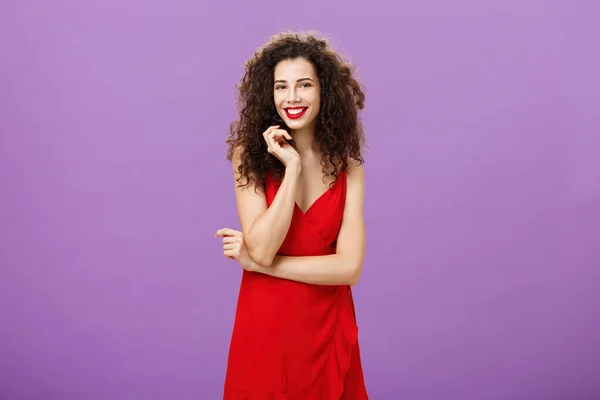 Πανέμορφη κυρία με κόκκινο αίσθημα ντροπαλός και αμήχανος που προσκαλείται για χορό στέκεται δειλό και θηλυκό πάνω από μωβ φόντο αγγίζοντας σκέλος σγουρά μαλλιά χαμογελώντας ευρέως και διασχίζουν το χέρι πάνω από το στήθος — Φωτογραφία Αρχείου
