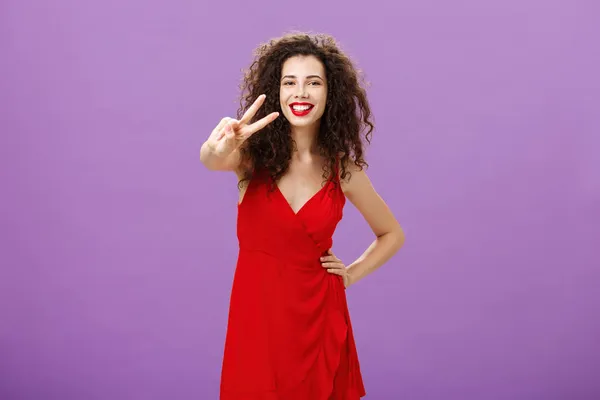 Studio girato di amichevole splendida ed elegante giovane donna europea con carino taglio di capelli ricci rossetto rosso e vestito che mostra gesto di pace alla fotocamera e sorridente ampiamente su sfondo viola — Foto Stock