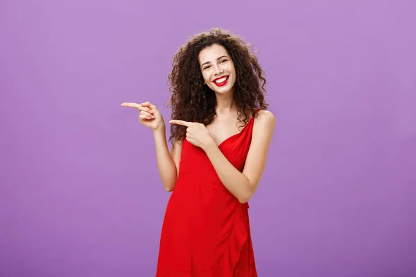 Κορίτσι προτείνει χώρο αντιγραφής για να δοκιμάσετε. Πορτρέτο της εμφανίσιμης κομψής καυκάσιας γυναίκας με σγουρά μαλλιά σε κομψό βραδινό κόκκινο φόρεμα που δείχνει αριστερά και χαμογελώντας καλώντας τους πελάτες πάνω από μωβ φόντο — Φωτογραφία Αρχείου