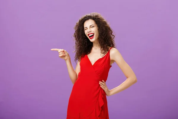 Χαρούμενη ανέμελη κομψή γυναίκα με σγουρό χτένισμα διασκεδάζοντας στο φοβερό πάρτι με κόκκινο πολυτελές φόρεμα κρατώντας το χέρι στη μέση κλείνει το μάτι και χαμογελώντας δείχνοντας ευρέως αριστερά, στέκεται πάνω από μωβ φόντο — Φωτογραφία Αρχείου