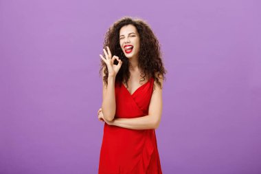 Havalı hevesli beyaz kadın, kıvırcık saç stili, neşeli bir şekilde dilini çıkartıyor ve onay işareti gösteriyor. Mor duvarın üzerinde tatmin olmuş ve kendinden emin bir şekilde poz veriyor.
