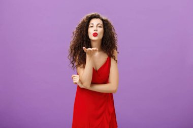 Tutkulu bir öpücük gönderen kadın umutsuz bir romantik. Zarif kırmızı elbise içinde, kıvırcık saç stili ve makyajlı. Gülümseyerek mwah üflüyor.