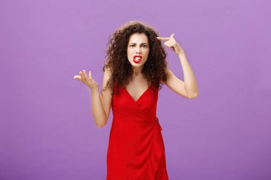 Outraged and pissed wife critisize dumb idea, outraged and fed up with stupid husband actions rolling finger near temple gesturing with raised palm, being angry and questioned over purple background