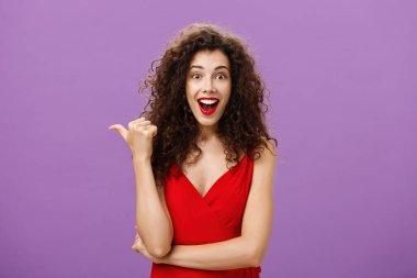 Kıvırcık saçlı, şaşkın ve şaşkın bir şekilde gülümseyen çekici ve güzel bir kadın. Başparmağını gösterip mor duvarın üzerinde kırmızı elbise içinde ilginç bir görüntü olup olmadığını soruyor.