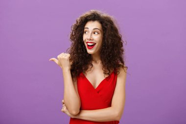 Kırmızılı, kıvırcık saçlı, gece makyajlı, şaşkın bakışlı, etkileyici bakışlı, hayranlık uyandıran ve mor duvarın üzerinde buluşurken arkadaşından soru soran tutkulu ve çekici bir kadın.