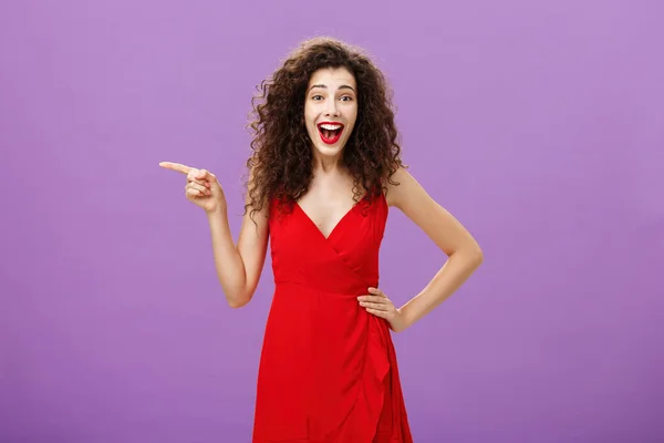 歓楽的で楽しいヨーロッパの女性の肖像で、赤い夜のドレスは遊び心と喜びから笑って左を指して紫色の背景の上にポーズを楽しんでいます — ストック写真