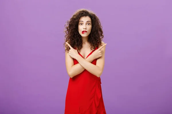Ενθουσιαστική αβέβαιη συναισθηματική κομψή γυναίκα σε κομψό βράδυ κόκκινο φόρεμα δάγκωμα χείλος αναζητούν ενθουσιασμένος δείχνει σε διαφορετικές κατευθύνσεις αριστερά και δεξιά είναι αβέβαιο και αμφίβολο στη λήψη απόφασης — Φωτογραφία Αρχείου