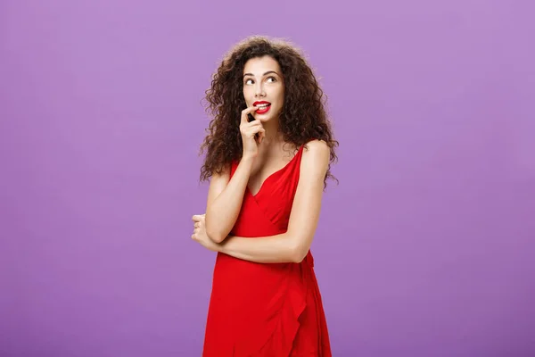 Ανόητη και συνεσταλμένη γοητευτική γυναίκα με σκούρα σγουρά μαλλιά σε κομψό βραδινό φόρεμα με κόκκινο κραγιόν που δαγκώνει το δάχτυλο που φαίνεται χαριτωμένο στην επάνω δεξιά γωνία στοχαστική απεικόνιση ή ονειρεύεται το επιθυμητό πράγμα — Φωτογραφία Αρχείου