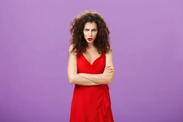 Kırmızılı, zarif kırmızı gece elbisesi giymiş, göğsünü göğsüne dayamış, öfkeyle alnının altından bakan ve eleştirilen kızgın bir kadının stüdyo fotoğrafı. — Stok fotoğraf
