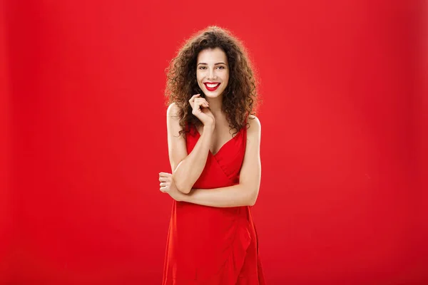 Παθιασμένη γοητευτική Ευρωπαία γυναίκα πάνω από το κόκκινο φόντο σε κομψό φόρεμα με σγουρό χτένισμα χαμογελαστό χαριτωμένο, θηλυκό παίζει με το σκέλος των μαλλιών στέκεται δειλό και ανόητο, μιλώντας στο πρόσωπο που θαυμάζει — Φωτογραφία Αρχείου