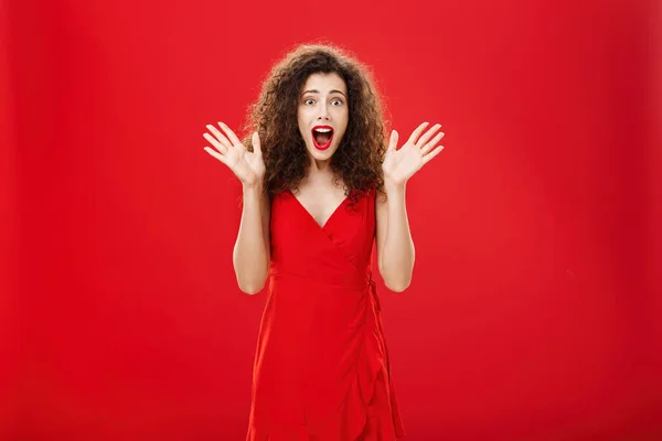 Apaixonada mulher adulta europeia espantada e emocionada em vestido de noite com penteado encaracolado gritando de deleite e surpresa acenando com as mãos levantadas e abrindo a boca de espanto sobre a parede vermelha — Fotografia de Stock