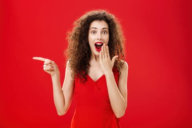 Mutlu, şaşkın ve keyifli, gece elbisesi içinde kıvırcık saçlı, ağzı açık, ağzı açık, ağzı açık, ağzı açık, solu işaret eden, kırmızı arka planda sorgulanmış ve heyecanlı bir kadın.