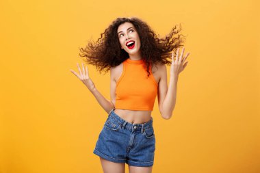 Aptal, pozitif ve enerjik, kıvırcık saçlı, kıvırcık saçlı, saçlarını savurup gülümseyen, sağ üst köşedeki hayalperest kadının portresi turuncu duvarın üzerinde mutlu poz veriyor.