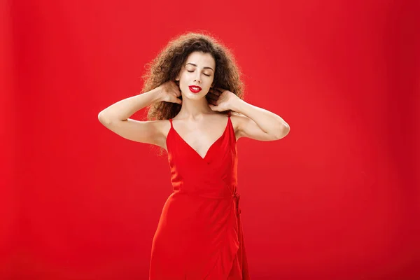 Εσωτερική λήψη της χαλαρής γυναίκας μηνυμάτων πίσω από το λαιμό με κλειστά μάτια και ήρεμη ξέγνοιαστη έκφραση στέκεται σε κομψό βραδινό φόρεμα με σγουρά χτένισμα κατά το κόκκινο φόντο ευχαριστημένος και κουρασμένος — Φωτογραφία Αρχείου