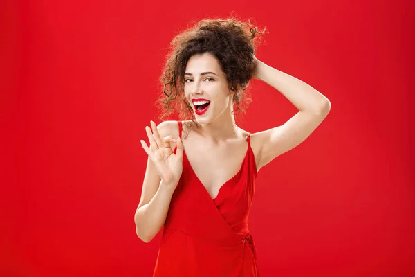 Retrato de sensual y sexy complacido despreocupado mujer adulta recogiendo el pelo en la espalda con la mano sonriendo alegremente y confiado mientras que muestra bien o gesto de aprobación sonriendo asegurando sobre fondo rojo —  Fotos de Stock