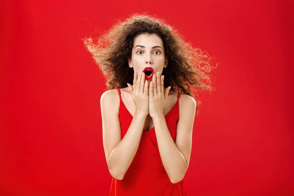 Atraktivní elegantní mladá žena v červených večerních šatech s kudrnaté vlasy mihotání na větru říká, wow je ohromen a vzrušený kryt otevřených úst z úžasu stojí nad červeným pozadím — Stock fotografie