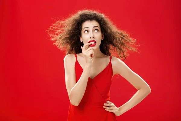 Cintura-up tiro de preocupado e preocupado inseguro bonito fêmea com penteado encaracolado no vestido de noite vermelho mordendo o dedo olhando perturbado ou questionado no canto superior direito com a mão na cintura — Fotografia de Stock