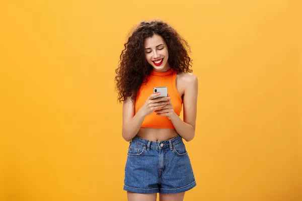 Girl spending time in internet texting friend messages via smartphone laughing while looking at device screen standing happy and upbeat over orange background in cropped top and denim shorts — Stock Photo, Image