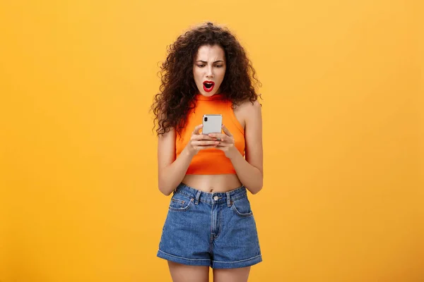 Woman being displeased and offended with low rate of photo in social network profile gasping from disappointment frowning and looking dissatisfied at smartphone screen standing over orange background