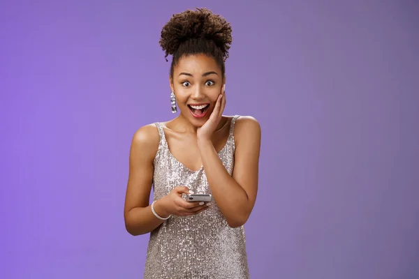 Surprised excited good-looking african american woman in silver glittering dress touch palm amazed widen eyes impressed drop jaw gasping holding smartphone astonished receive good news phone