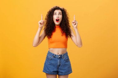 Şaşırmış, duygusal ve karizmatik genç, kıvırcık saçlı, kırmızı rujlu bir kadının portresi turuncu arka planda etkilenmiş ve hayretler içinde kalmış.