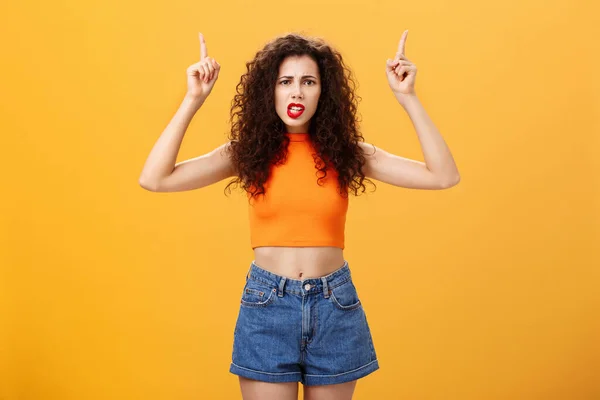 Indoor shot of displeased female client with curly stylish hairstyle red lipstick in cool cropped top pointing up demanding explanation with confused and dissatisfied expression over orange background