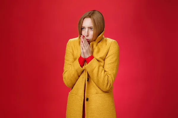 黄色の光のコートを身に着けている強烈な震えかわいい赤毛の女性のスタジオショット手をこすり、暖かい息を手のひらで吹いて、暖かい感じの寒さ、赤の背景に冷たい風の天気の間に凍結 — ストック写真